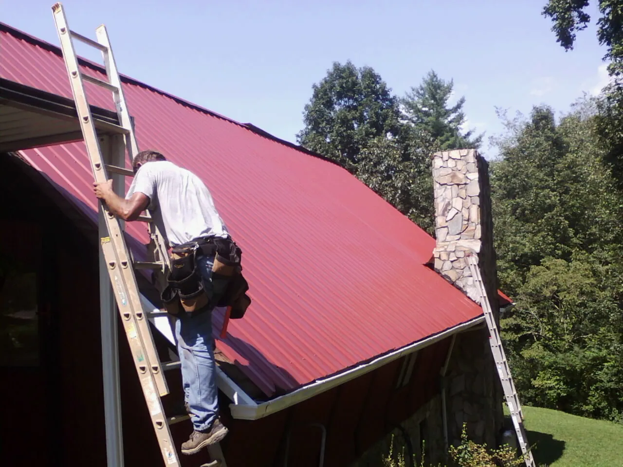 Roofer on a ladder beside a red metal roof and stone chimney — Soffit & Fascia Repair by Roofing Brothers in French Lick